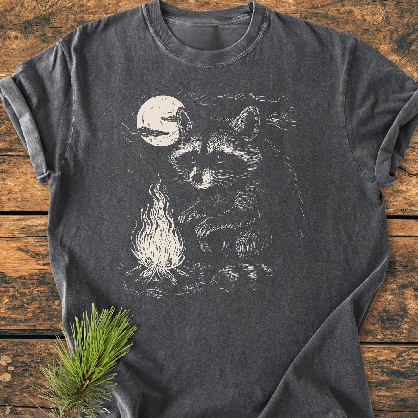 1409 Moonlit Campfire Raccoon Base T-shirt featuring a raccoon by a campfire illustration, laid flat on wood with a pine branch, showcasing its relaxed fit and durable stitching.