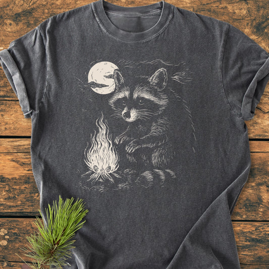 1409 Moonlit Campfire Raccoon Base T-shirt featuring a raccoon by a campfire illustration, laid flat on wood with a pine branch, showcasing its relaxed fit and durable stitching.