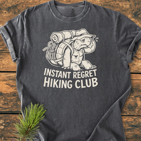 Charcoal-gray T-shirt with a cartoon of an overloaded turtle and INSTANT REGRET HIKING CLUB slogan, showcasing a relaxed, vintage look with a pine sprig accent on a wooden surface.