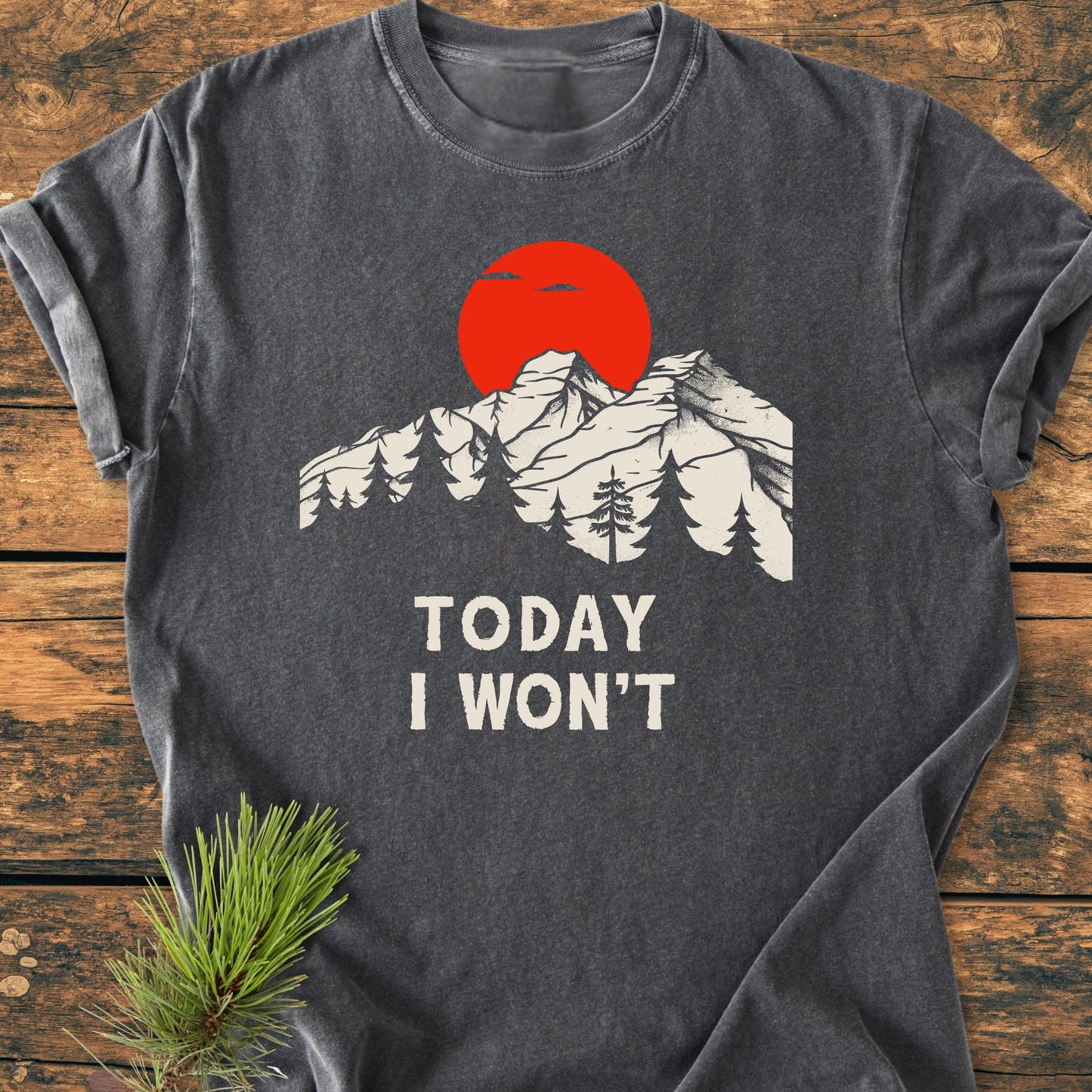 Charcoal-gray T-shirt, 1423 Today I Won't, features a mountain scene and red sun. Laid on wood, with pine sprig accentuating its soft, vintage wash appeal for everyday wear.