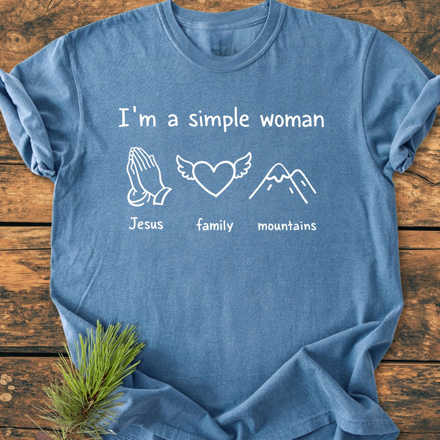 Blue T-shirt with “I’m a Simple Woman” in white, featuring icons of praying hands, a heart with wings, and mountains. Laid flat on wood with pine sprig, showcasing vintage wash texture.