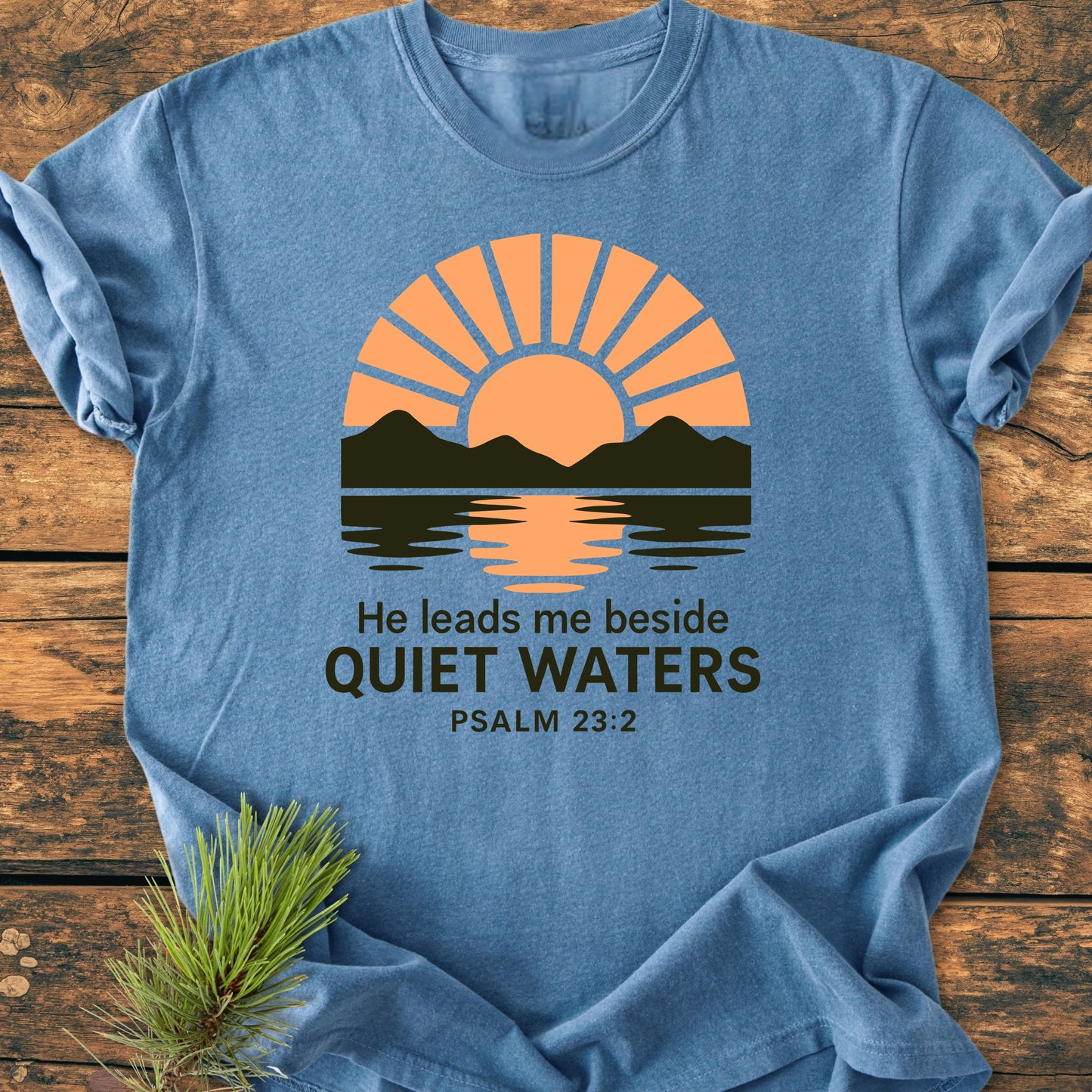 Heather-blue T-shirt titled 1424 He Leads Me, featuring an orange sun and mountain graphic with He leads me beside QUIET WATERS PSALM 23:2, showcasing a vintage wash and relaxed fit.