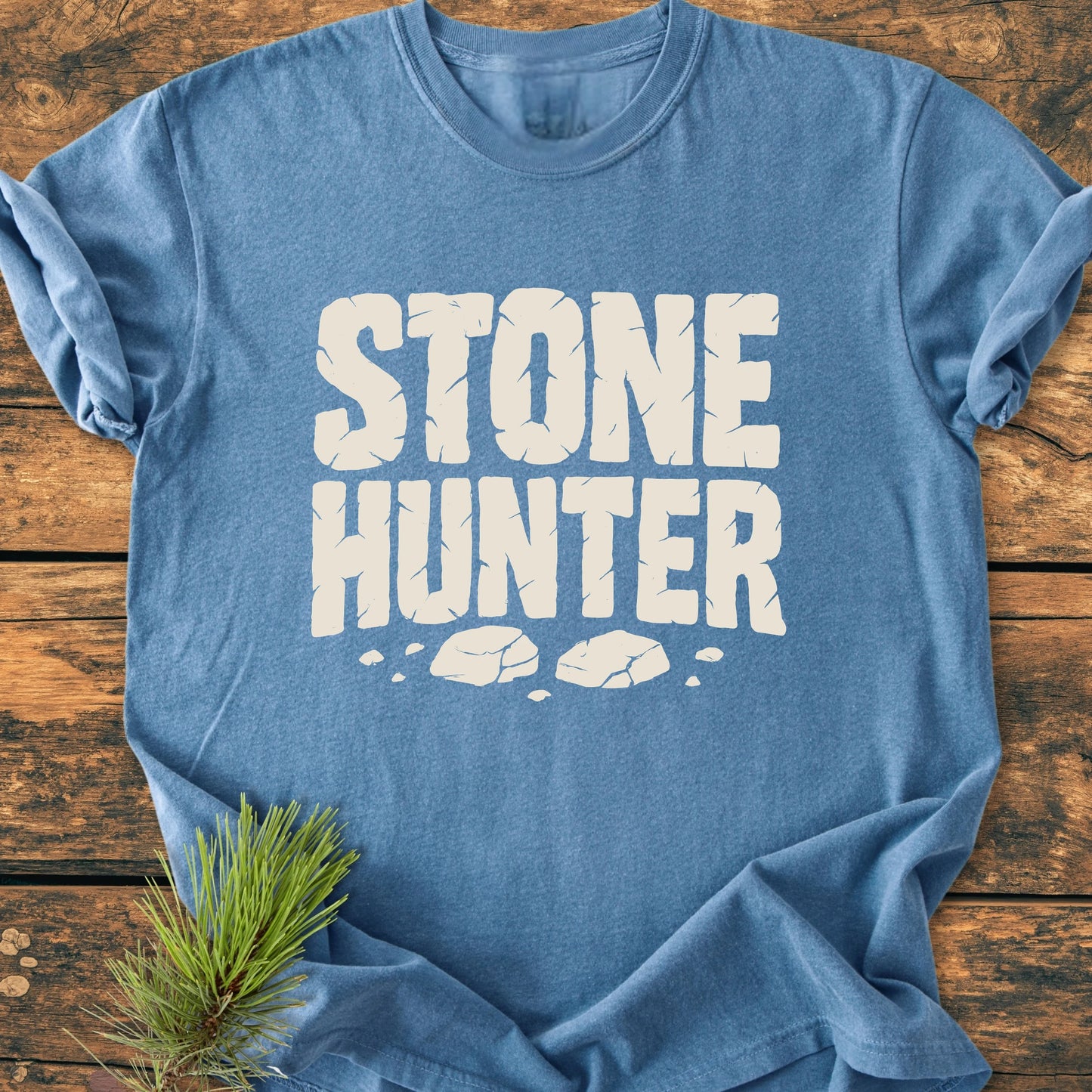 Heather-blue T-shirt featuring STONE HUNTER in cracked-stone style text, with rock illustrations below. A pine sprig adorns the tee, highlighting its vintage wash and relaxed fit.