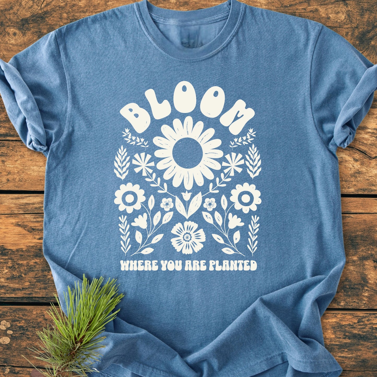 Heather-blue T-shirt with Bloom Where You Are Planted slogan and floral design, laid flat on wood, showcasing its vintage wash and relaxed fit, perfect for everyday wear from Trails & Tees.