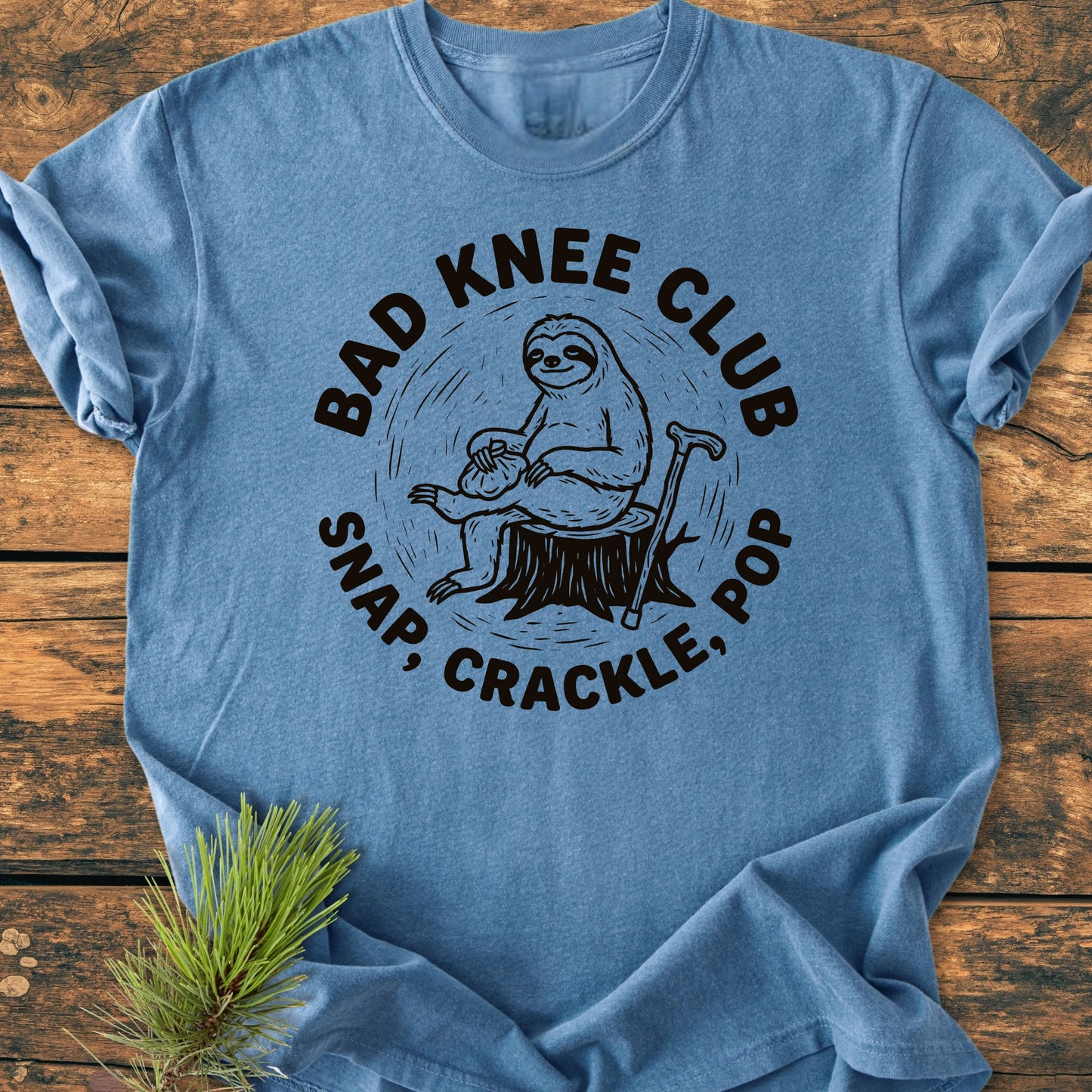 Blue T-shirt featuring a cartoon sloth with an ice-pack on its knee, sitting on a stump. Encircling text reads “BAD KNEE CLUB” and “SNAP, CRACKLE, POP”, conveying a playful, relaxed vibe.