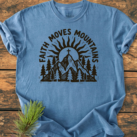 Heathered-blue 1409 Faith Moves Mountains T-shirt featuring a black screen-printed mountain and pine tree scene, laid on wood, showcasing sunrays and a pine sprig accent.