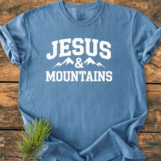 Heather-blue T-shirt with “JESUS & MOUNTAINS” in white, featuring mountain peaks graphic. Laid on wooden surface, accompanied by a pine branch, showcasing a vintage, relaxed fit ideal for outdoor adventures.