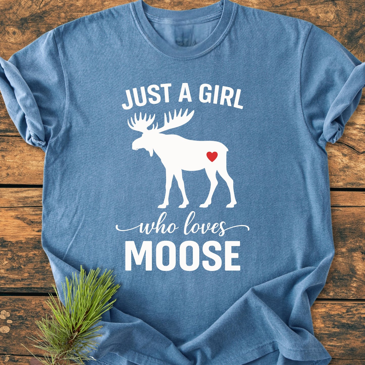 Blue T-shirt with “1437 Just A Girl Who Loves Moose,” featuring a white moose silhouette with a red heart, laid on wood with pine sprig, showcasing vintage, soft-washed feel.