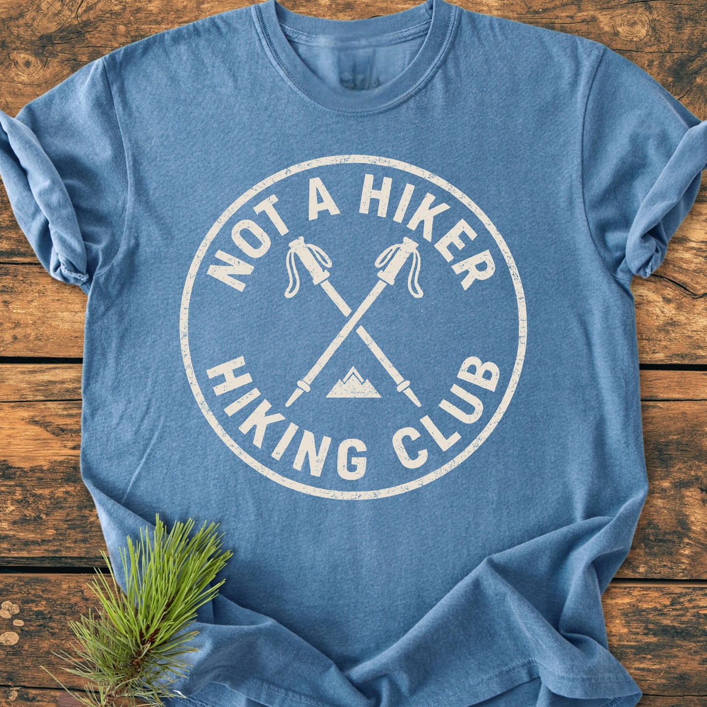 Heather-blue 1426 Not A Hiker T-shirt features a white distressed circle design with hiking poles and mountain, laid on wood with a pine sprig, highlighting its vintage casual appeal.