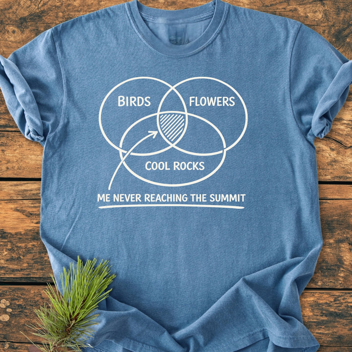 Heather-blue T-shirt titled 1416 Easily Distracted Hiker features a white Venn diagram with overlapping circles labeled “BIRDS,” “FLOWERS,” and “COOL ROCKS,” and a humorous caption, laid on a wooden surface.