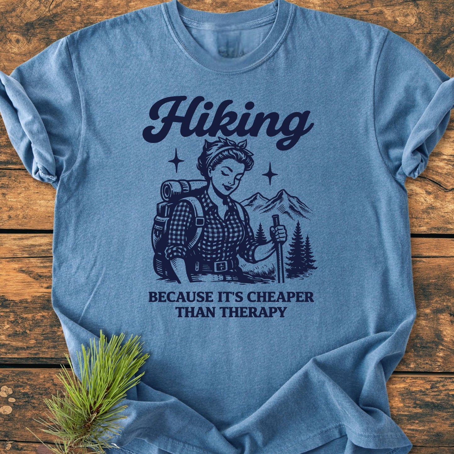 The 1407 Hiking Cheaper Than Therapy Base T-shirt features a vintage illustration of a woman hiking, with mountains and trees, highlighting its comfort and durability on a wooden backdrop.