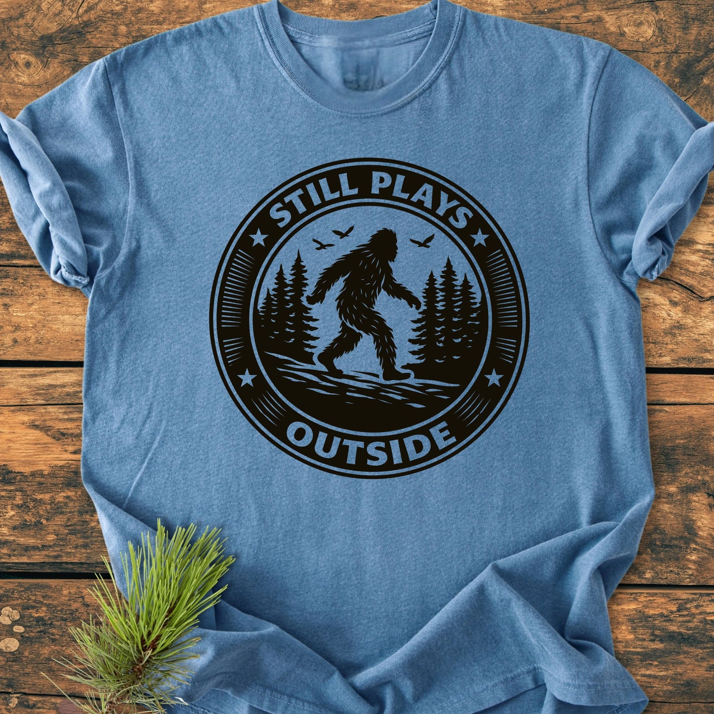Heathered-blue T-shirt featuring 1430 Still Plays Outside Bigfoot design: a black circular graphic with Bigfoot silhouette, pine trees, and birds, laid on rustic wood with a pine sprig nearby.