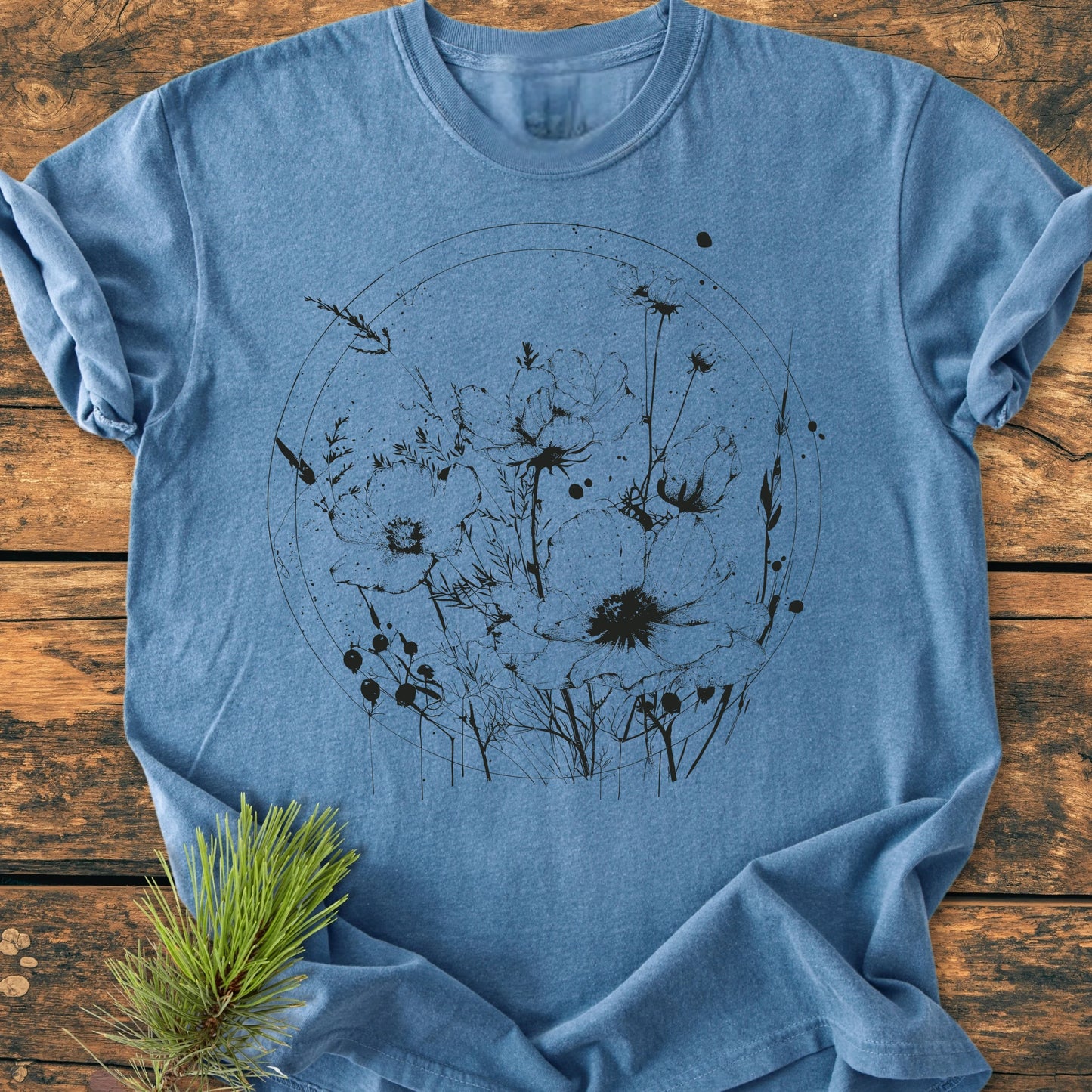 Heather-blue 391 Wildflower Circle T-shirt with black wildflower line-art, laid on wood. Features 100% ring-spun cotton, relaxed fit, and durable stitching, emphasizing comfort and nature-inspired style.