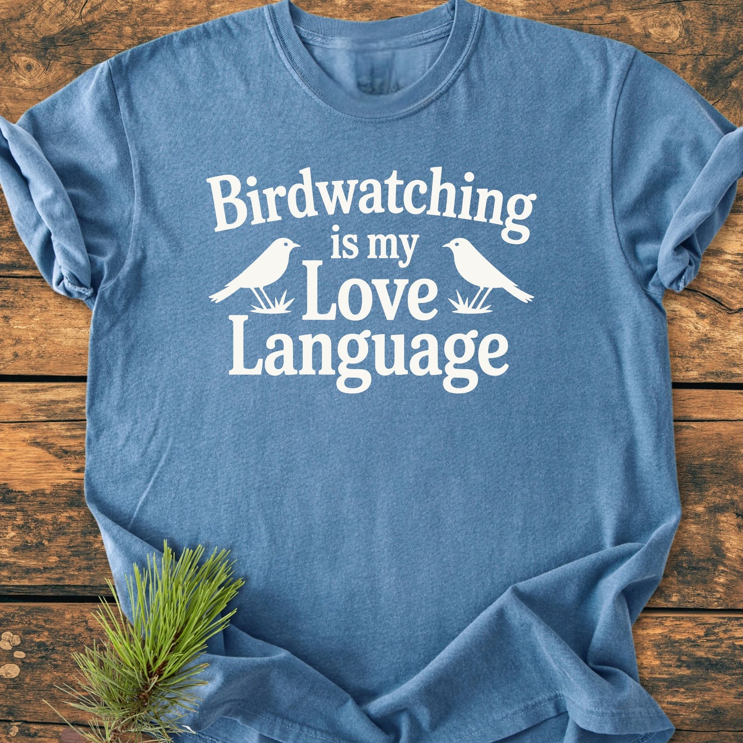 Blue T-shirt with Birdwatching is my Love Language printed in white, featuring bird silhouettes. Displayed flat with a pine twig under the edge. Product: 1428 Birdwatching Love Language.