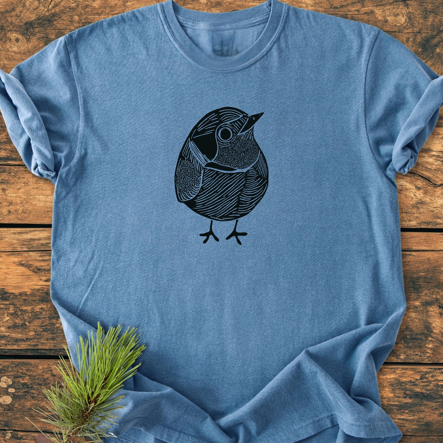 Soft-washed blue tee, 1433 Little Robin, featuring a sketchy bird line-drawing, laid on rustic wood. Styled with a pine twig, embodies vintage comfort and relaxed unisex fit from Trails & Tees.