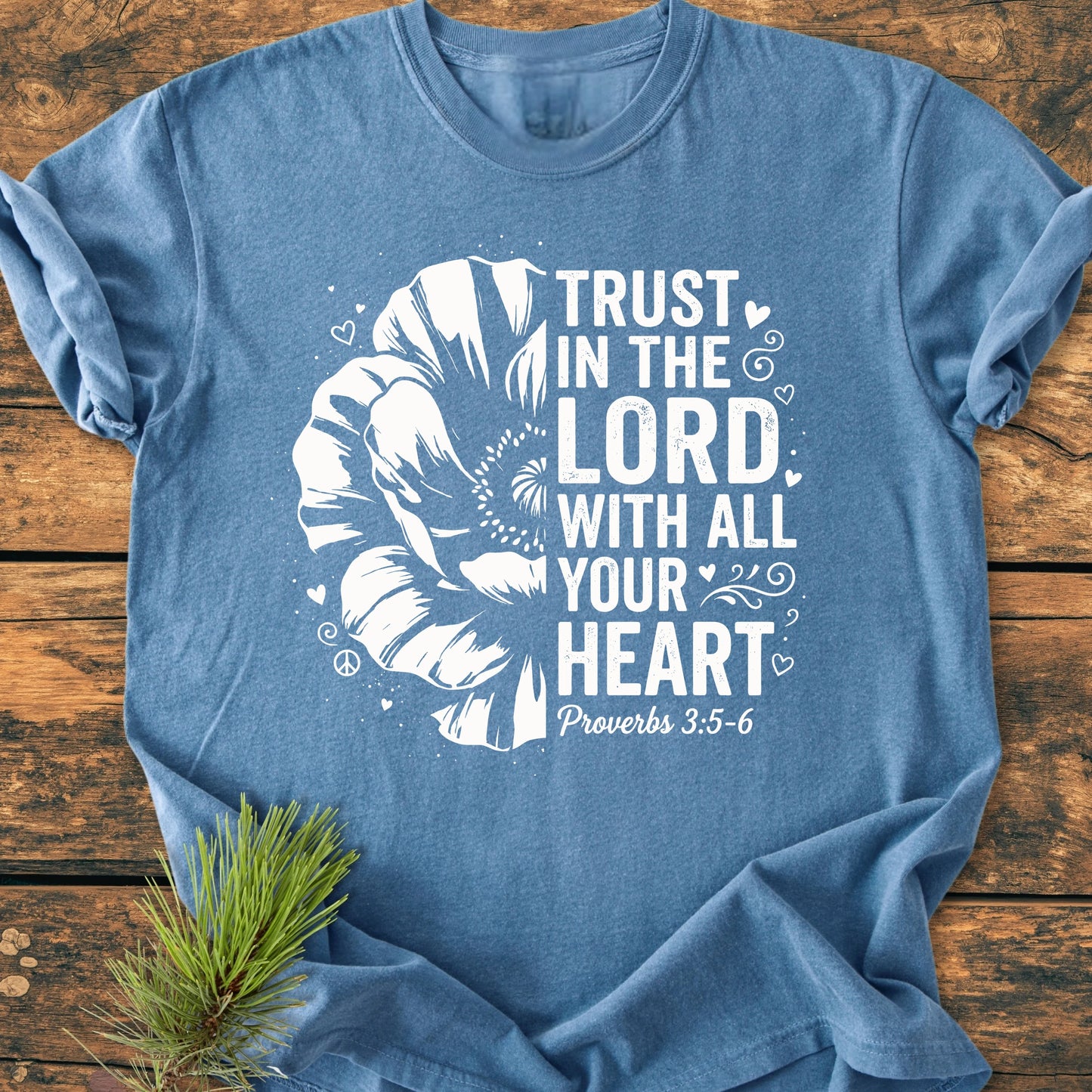 Trust In The Lord - Vintage Wash Tee: A heathered blue T-shirt with a white flower graphic and Bible verse. Lays flat on wood, accompanied by a small pine sprig.