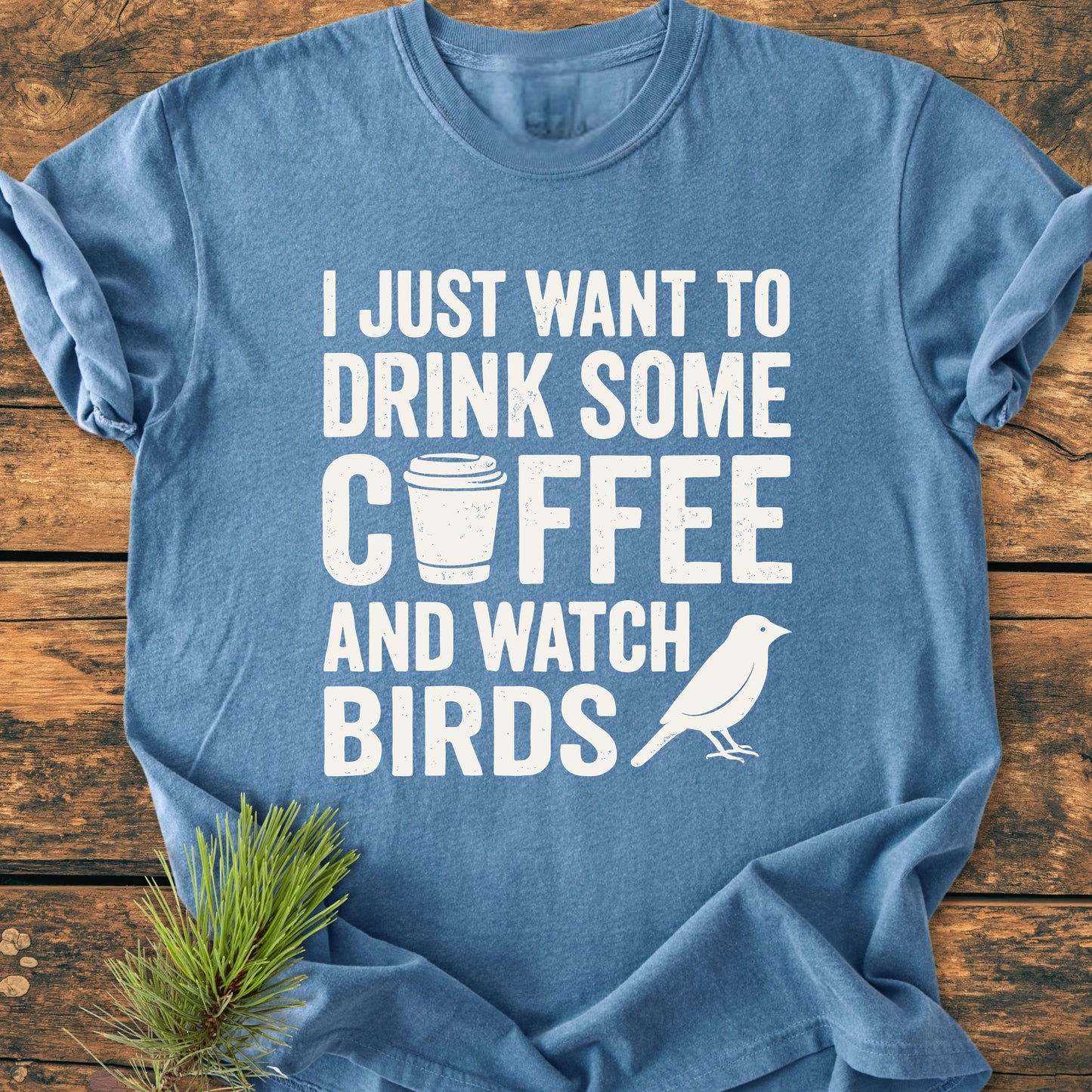 Soft-blue 1405 Drink Coffee And Watch Birds T-shirt on wood, featuring bold white text with coffee cup and bird icons, enhanced by a pine sprig, highlighting its cozy, durable design.