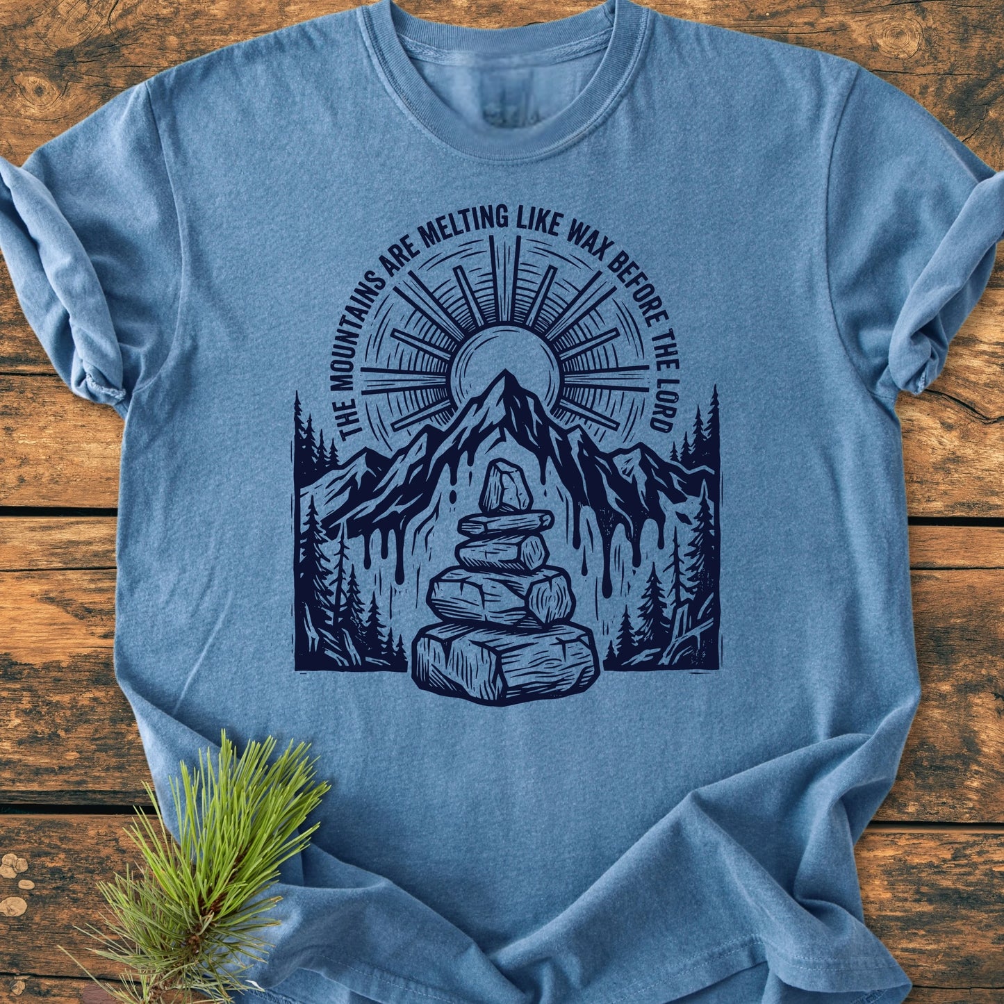 Light-blue T-shirt titled 1414 Mountains Melting Like Wax, featuring a mountain illustration with melting sides, evergreen trees, cairn, and sunburst, laid on rustic wood with a pine sprig beside.