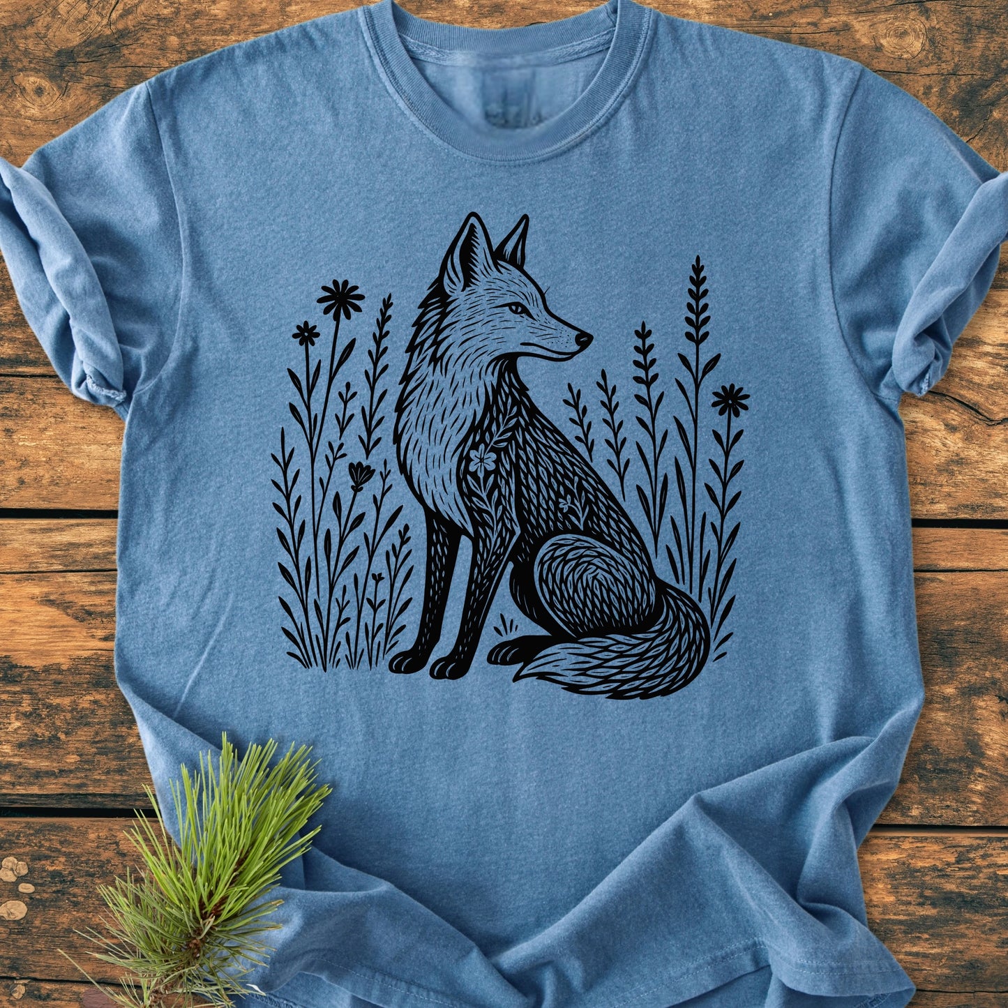 The 225 Wildflower Fox DARK T-shirt features a bold fox illustration amid wildflowers on a soft-blue, short-sleeve tee, styled with a pine sprig on a rustic wooden surface.