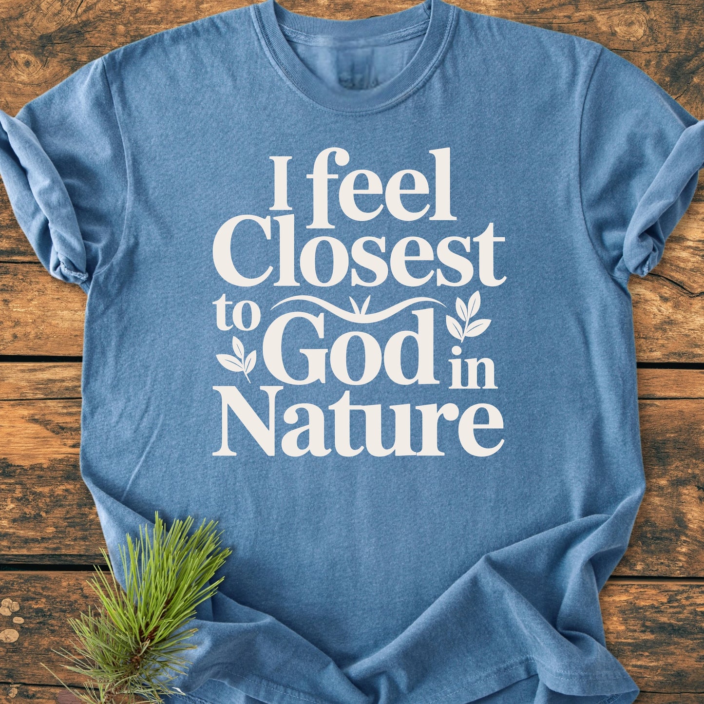 Heather-blue 1427 Closest To God - Vintage Wash Tee laid flat, featuring bold white text and leaf accents, on a rustic wooden surface with a pine sprig beside it.