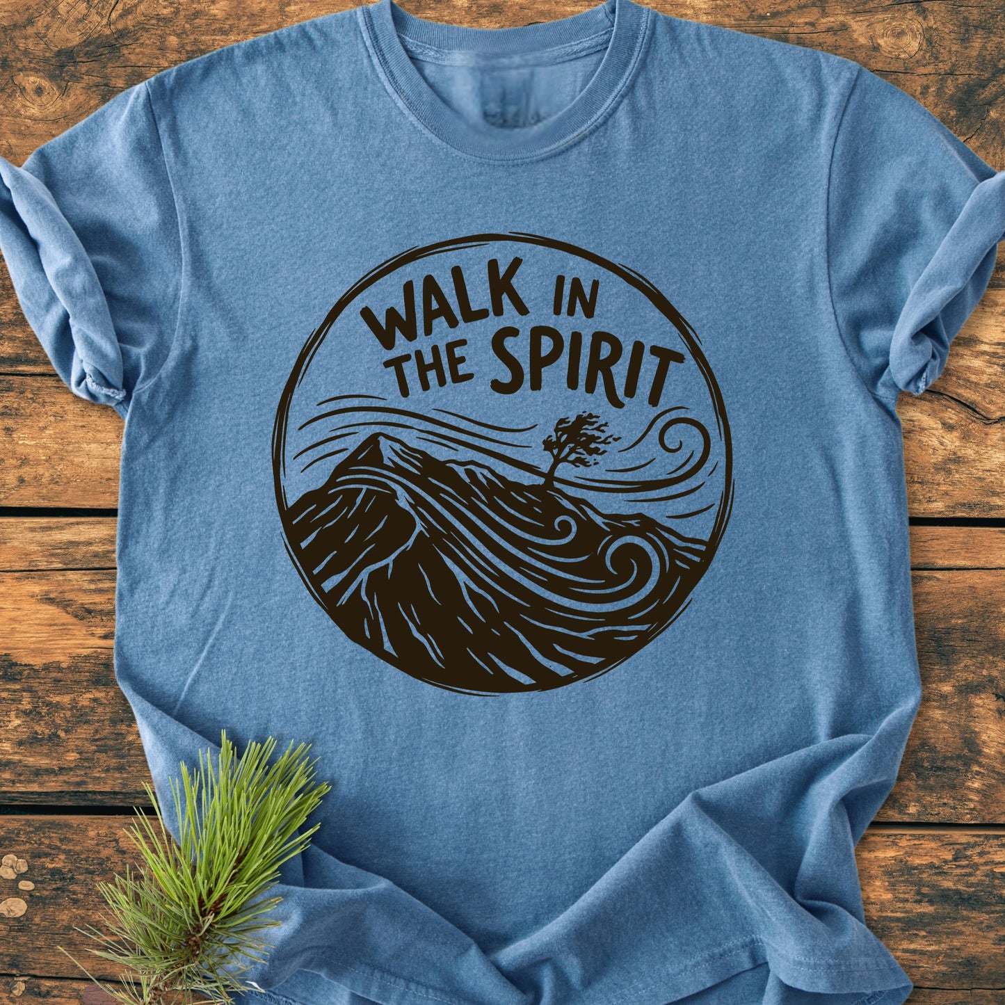 Heather-blue T-shirt featuring the 1410 Walk In The Spirit emblem with a mountain, windswept tree, and swirling lines, laid flat on wood beside a pine sprig.