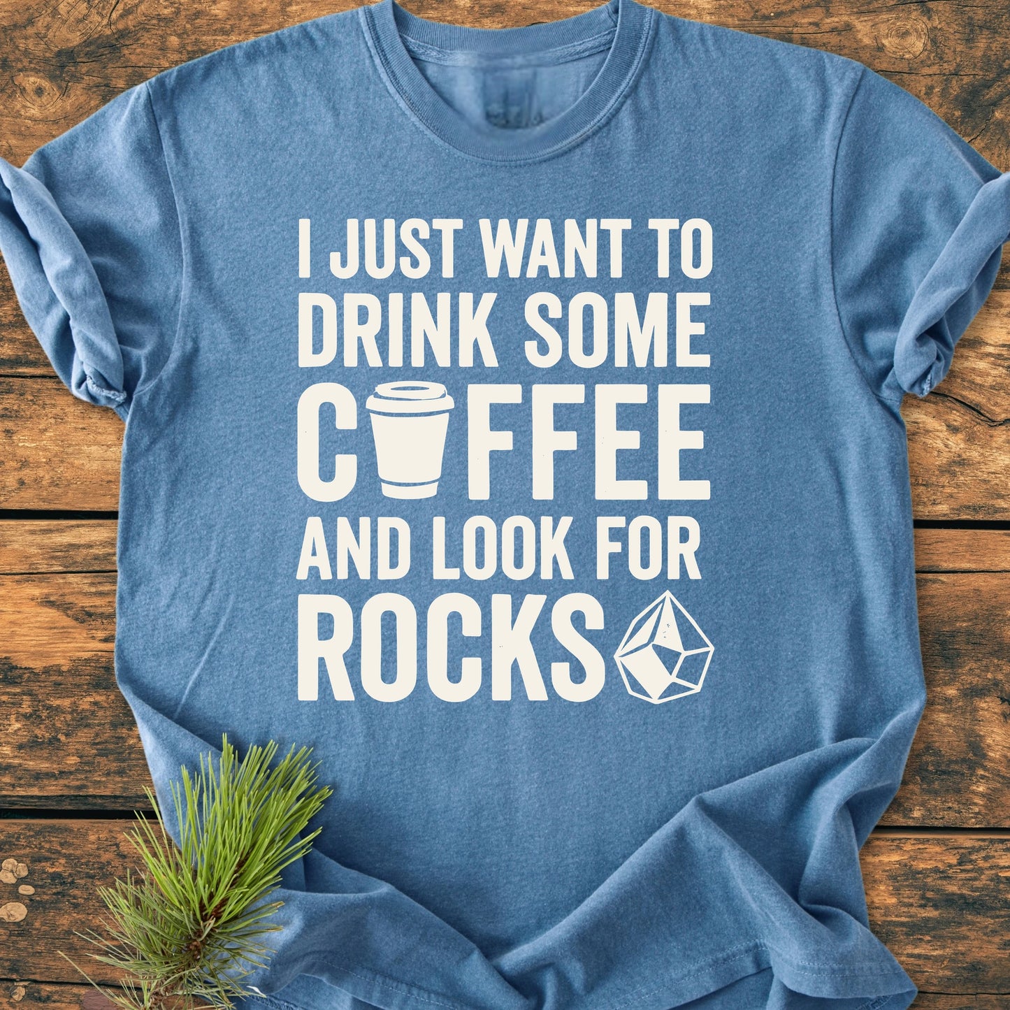 Heathered blue 1404 Drink Coffee And Look For Rocks Base T-shirt on wood, featuring bold white text with coffee cup icon and gem illustration, conveying a cozy, durable, and relaxed fit.