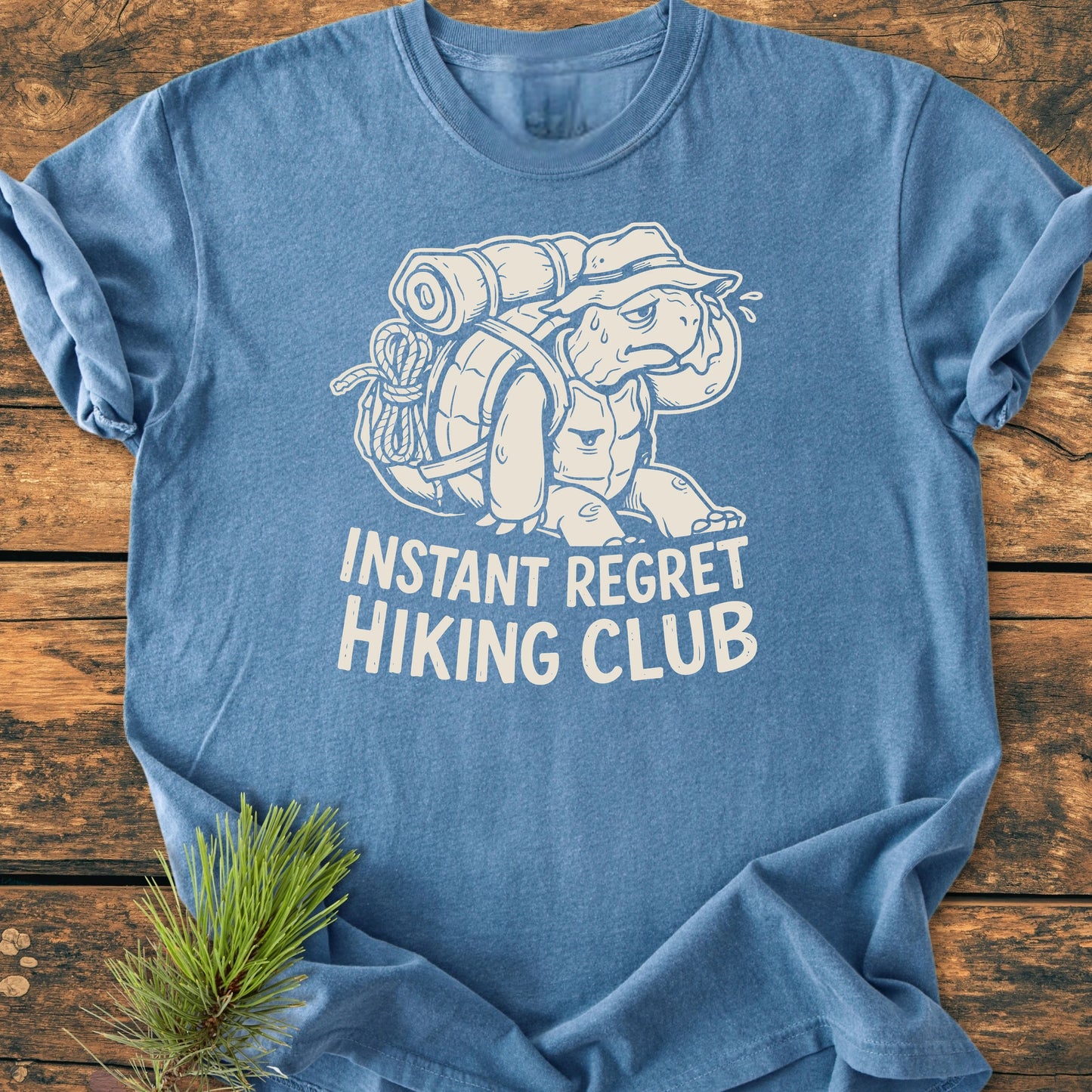 Heather-blue T-shirt with cartoon tortoise in a bucket hat and hiking pack, labeled INSTANT REGRET HIKING CLUB, laid out on wood. Vintage Wash Tee, relaxed fit, ideal for everyday wear.
