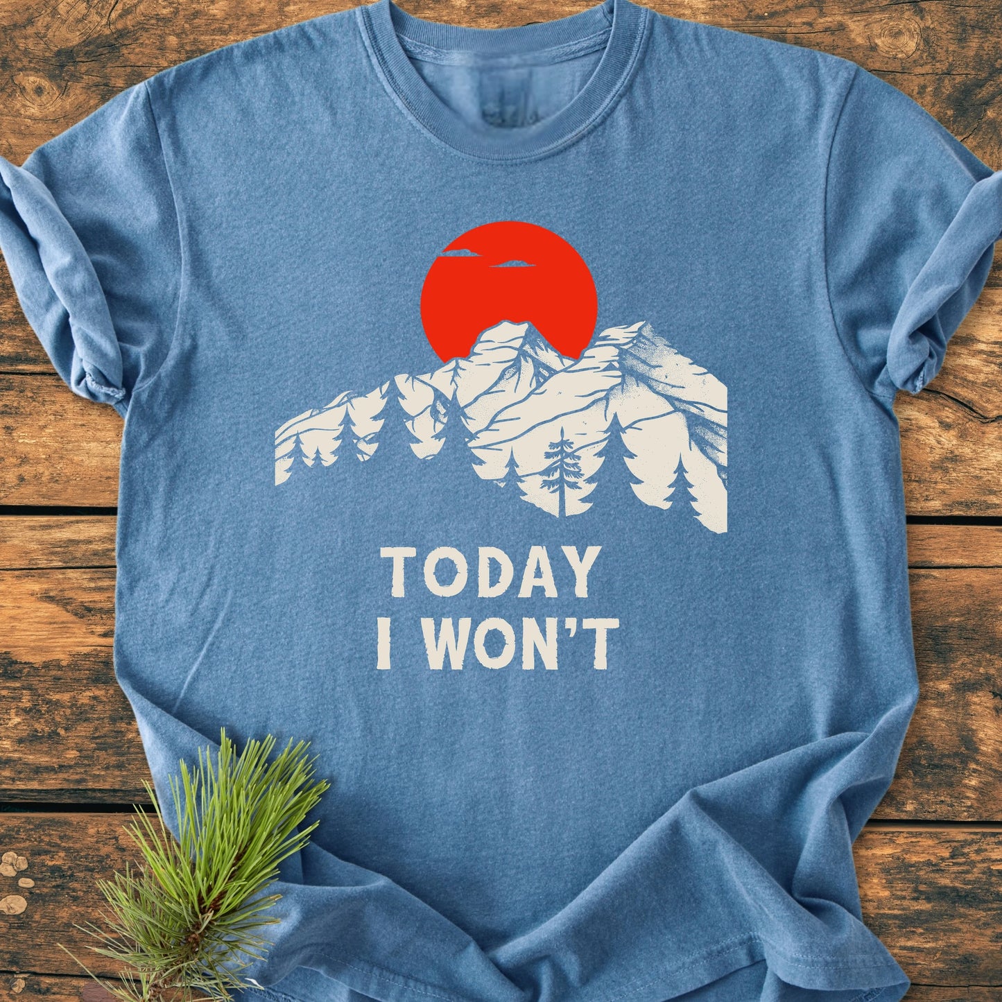 Heather-blue T-shirt titled 1423 Today I Won't features white mountain peaks, pine trees, and a red sun graphic, laid on wood with a pine sprig underneath, capturing a relaxed, vintage style.