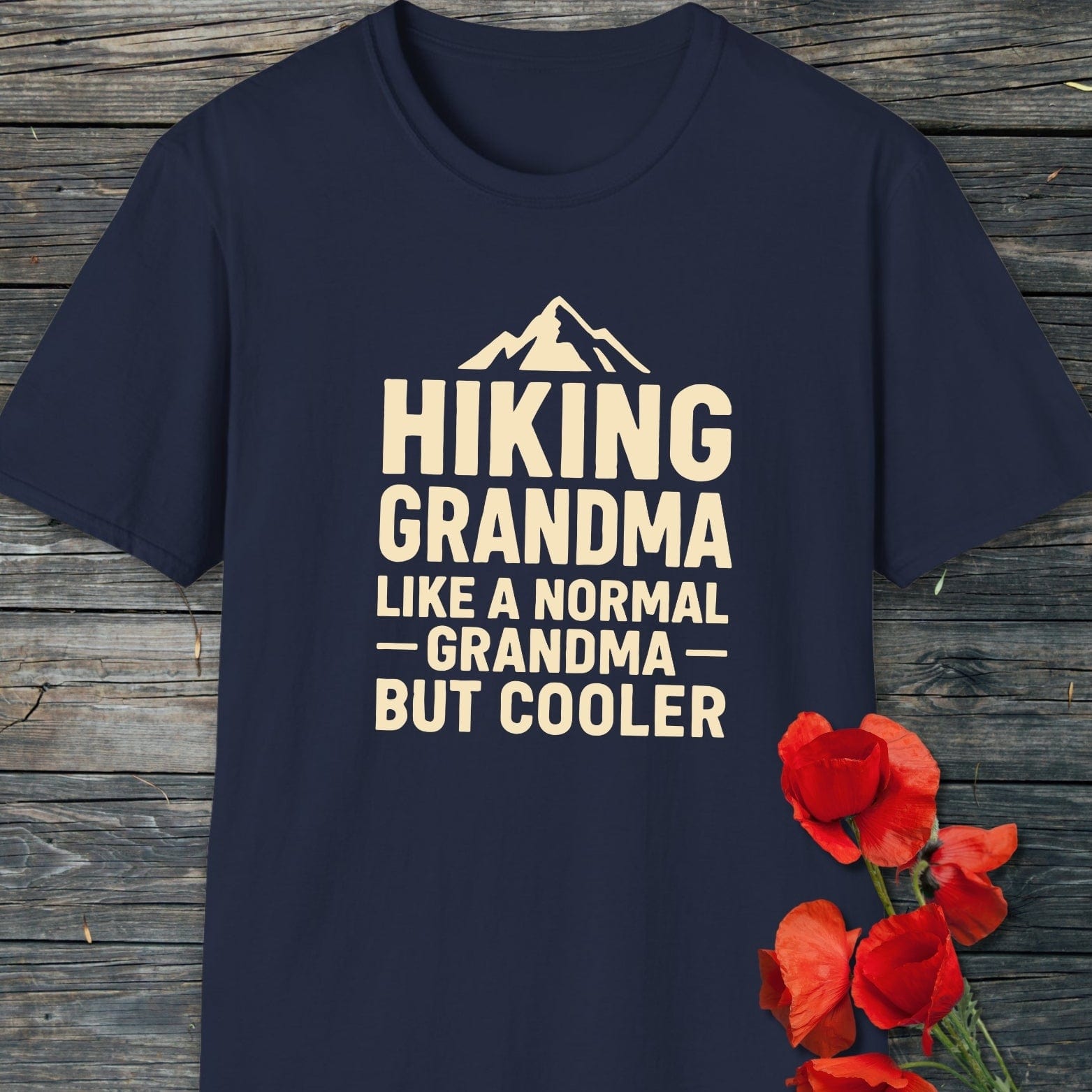Navy-blue Hiking Grandma T-shirt with mountain graphic, laid flat on wood, showcasing its soft, durable cotton fabric; accentuated by red flowers for decoration.