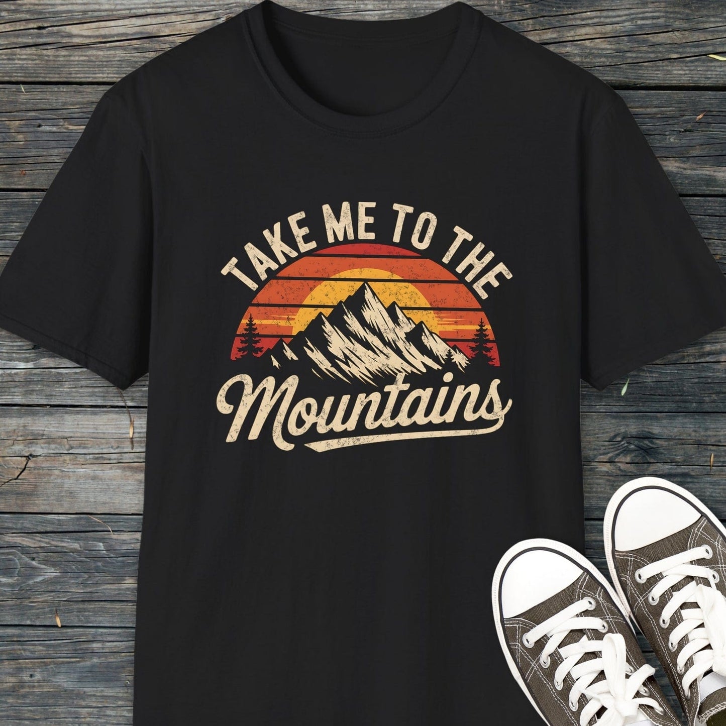 Take Me To The Mountains T-Shirt with vintage sunset graphic, laid on wood with sneakers. Made of soft, lightweight cotton for comfort and adventure.