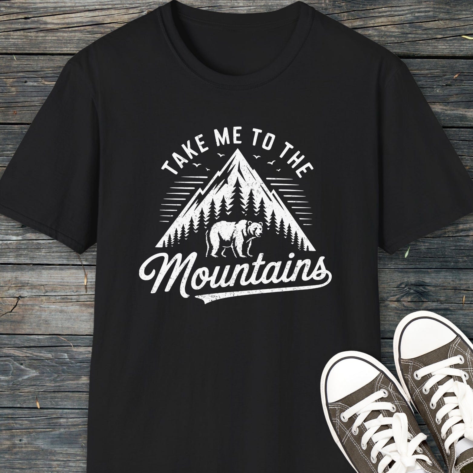 Take Me To The Mountains T-Shirt with vintage mountain graphic, pine trees, and bear silhouette, paired with canvas sneakers, highlighting adventure-ready comfort and unisex fit.