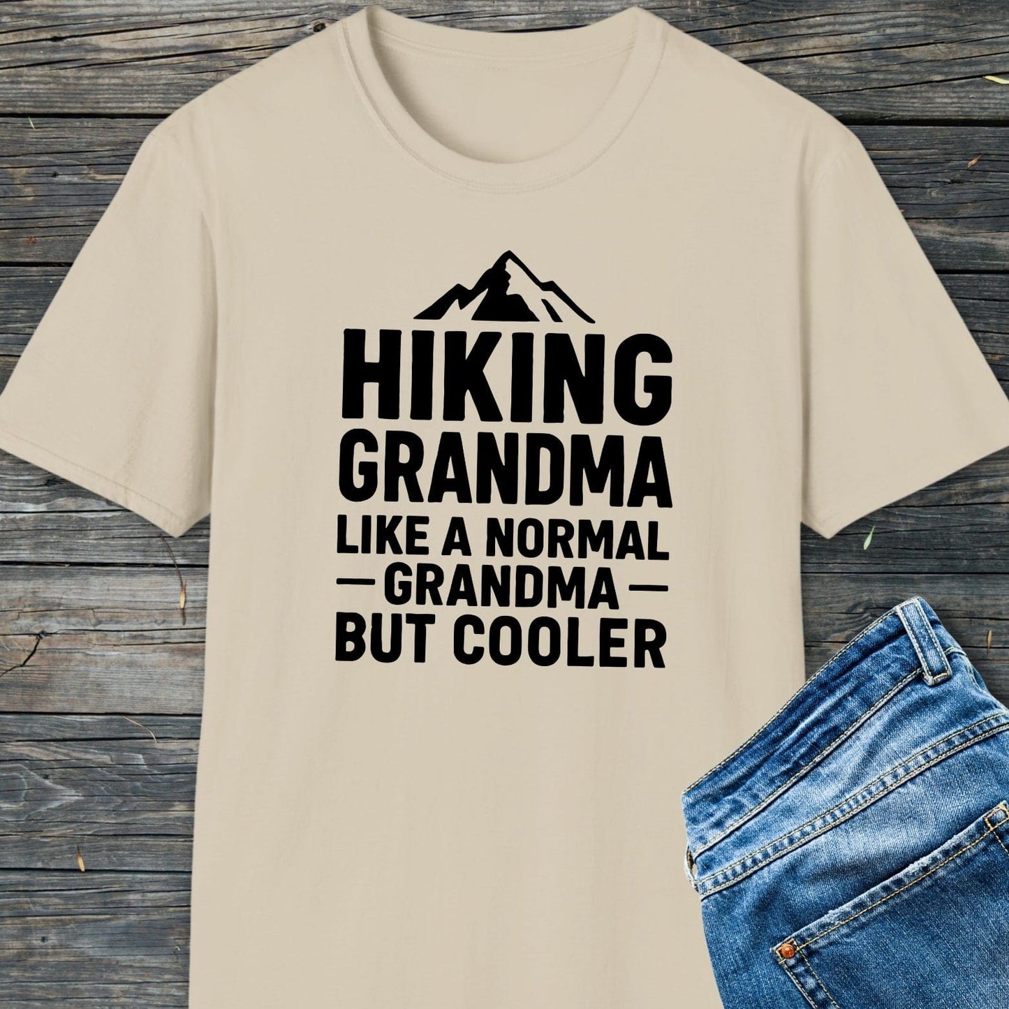 Hiking Grandma T-Shirt featuring a mountain graphic and bold text, laid beside blue jeans, crafted from soft ring-spun cotton for comfort and durability.
