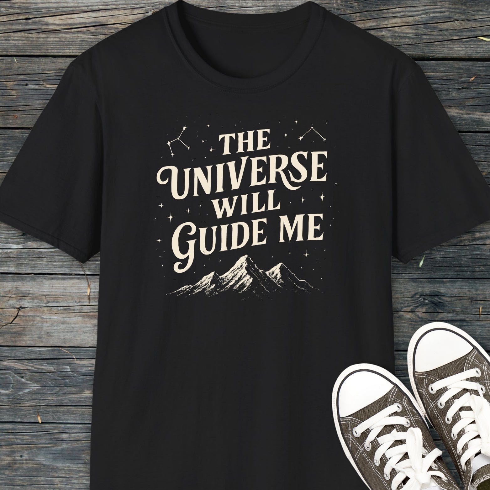 The Universe Guides Me T-Shirt featuring a cream graphic with stars, constellations, and mountains, laid on wood, with partial view of black canvas sneakers.