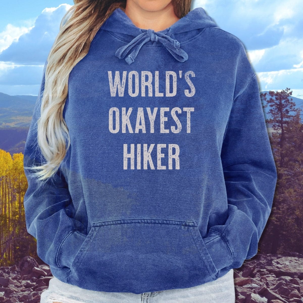 Person wearing a World's Okayest Hiker Comfort Colors Hoodie with hands in kangaroo pocket, standing outdoors with trees and hills in the background.