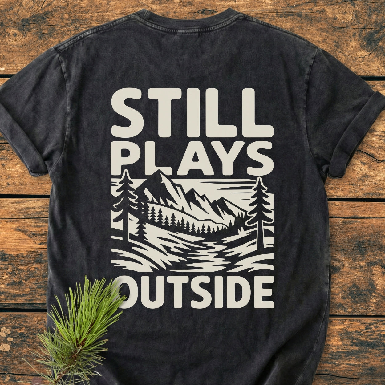 Still Plays Outside BACK PRINT - Vintage Wash Tee featuring a stylized mountain landscape graphic on a charcoal short-sleeve t-shirt, laid flat on wood with a pine sprig beside it.