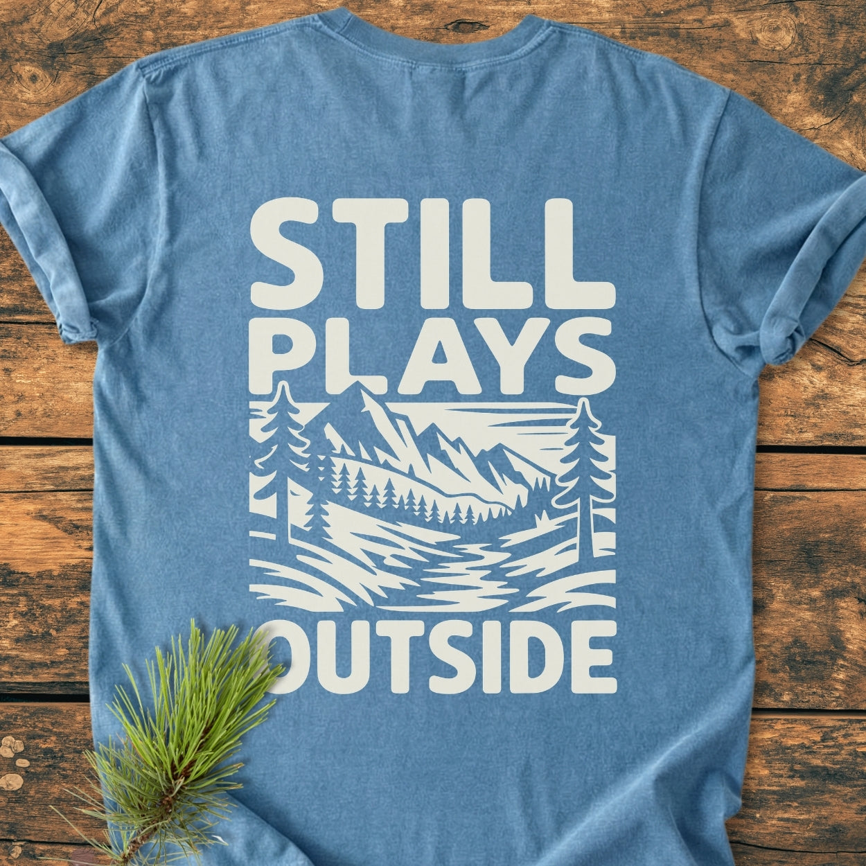 Still Plays Outside BACK PRINT - Vintage Wash Tee on wooden surface, showcasing a white mountain and forest scene design with slogan. Pine needles accent the shirt, highlighting its relaxed, durable, and cozy fit.