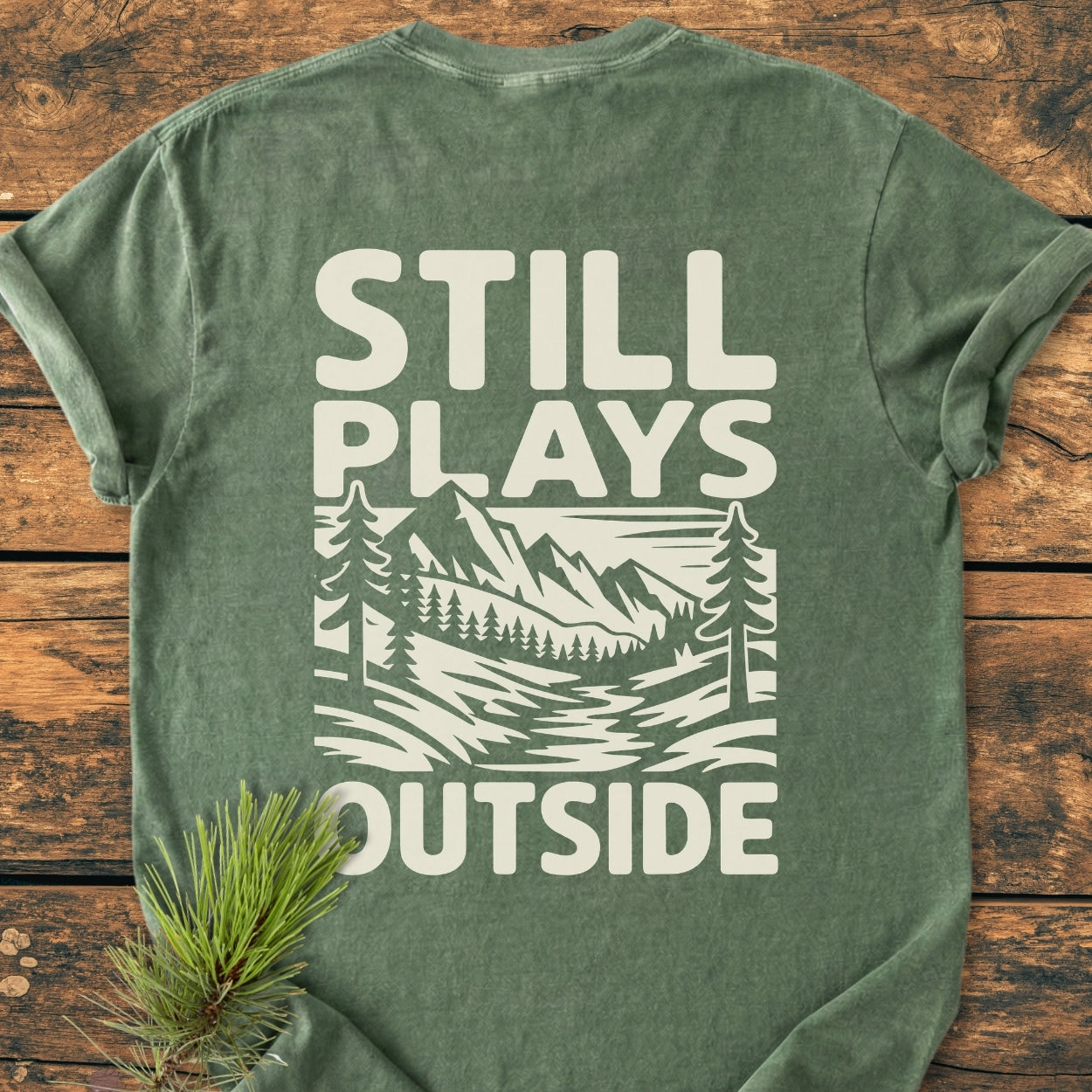 Still Plays Outside BACK PRINT - Vintage Wash Tee featuring a stylized outdoor scene with mountains and trees on a faded-green T-shirt, laid flat with a pine sprig accent on a wooden surface.