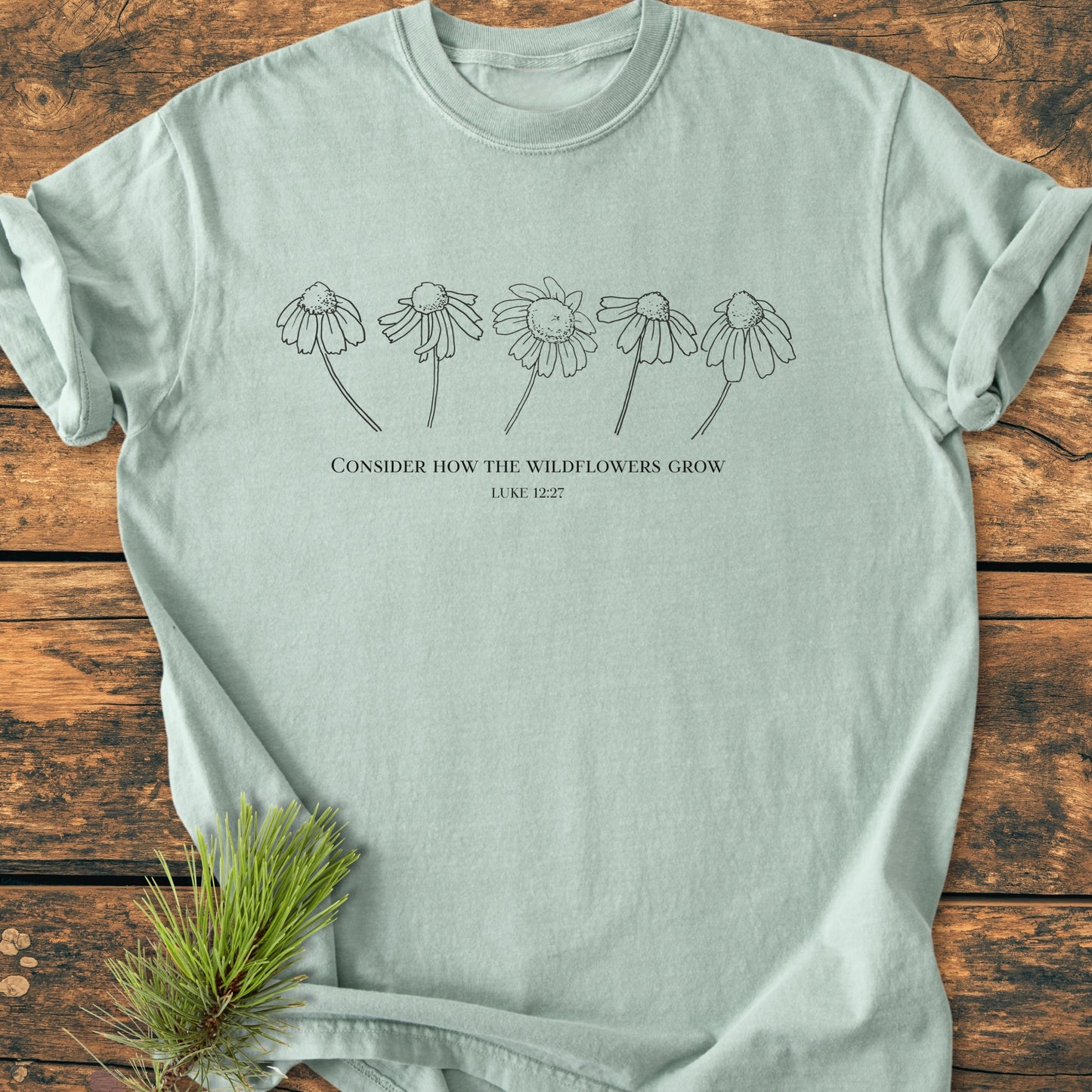 Consider How The Wildflowers Grow - Vintage Wash Tee: Mint-green cotton T-shirt featuring minimalist wildflower line art and biblical quote, styled on a wooden surface with a pine sprig accent.