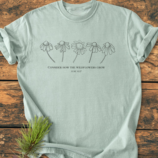 Consider How The Wildflowers Grow - Vintage Wash Tee: Mint-green cotton T-shirt featuring minimalist wildflower line art and biblical quote, styled on a wooden surface with a pine sprig accent.