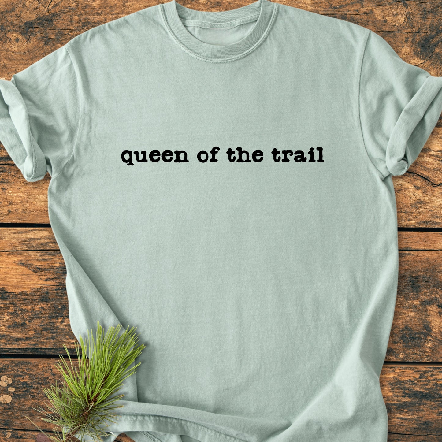 Queen Of The Trail - Vintage Wash Tee, a relaxed-fit mint crew-neck with bold black lettering, displayed on rustic wood, accented by a small pine branch for an outdoorsy touch.