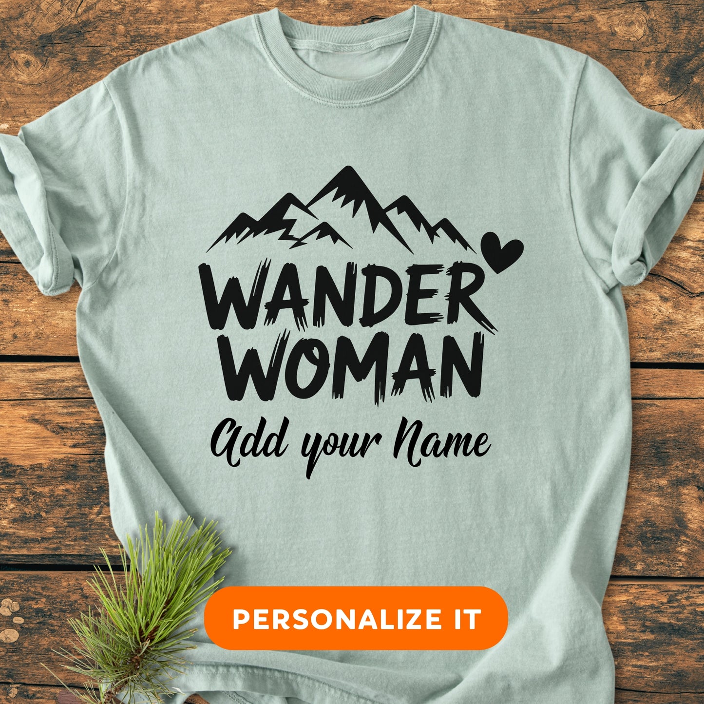 Wander Woman | Custom Name | Vintage Wash Tee featuring a black mountain graphic, heart, and personalization option, laid flat on wood with a pine sprig and PERSONALIZE IT button graphic below.