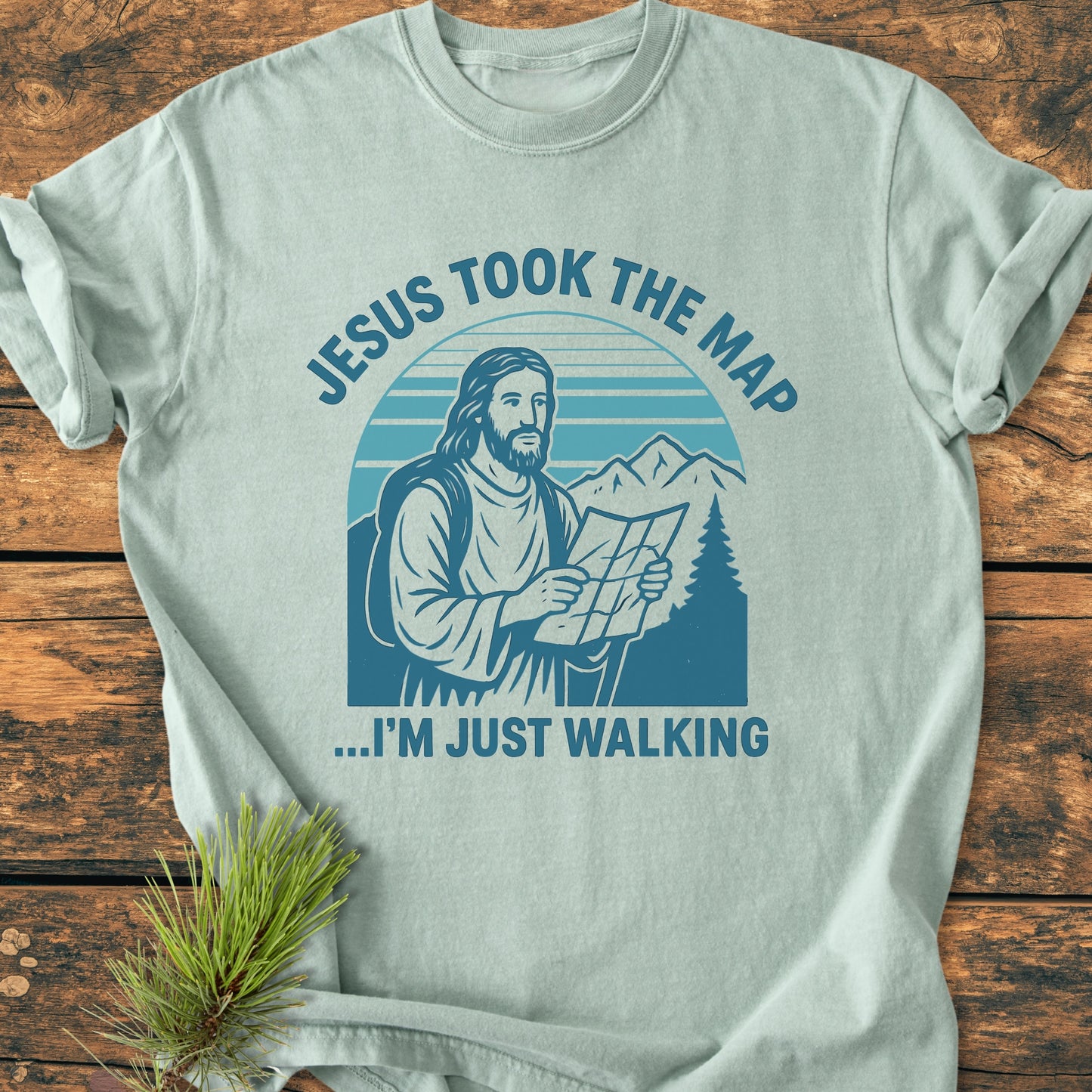 Jesus Took The Map - Vintage Wash Tee features a line drawing of Jesus with a backpack and map, set against mountains and trees, on a pale green T-shirt, enhancing a relaxed, comfortable style.