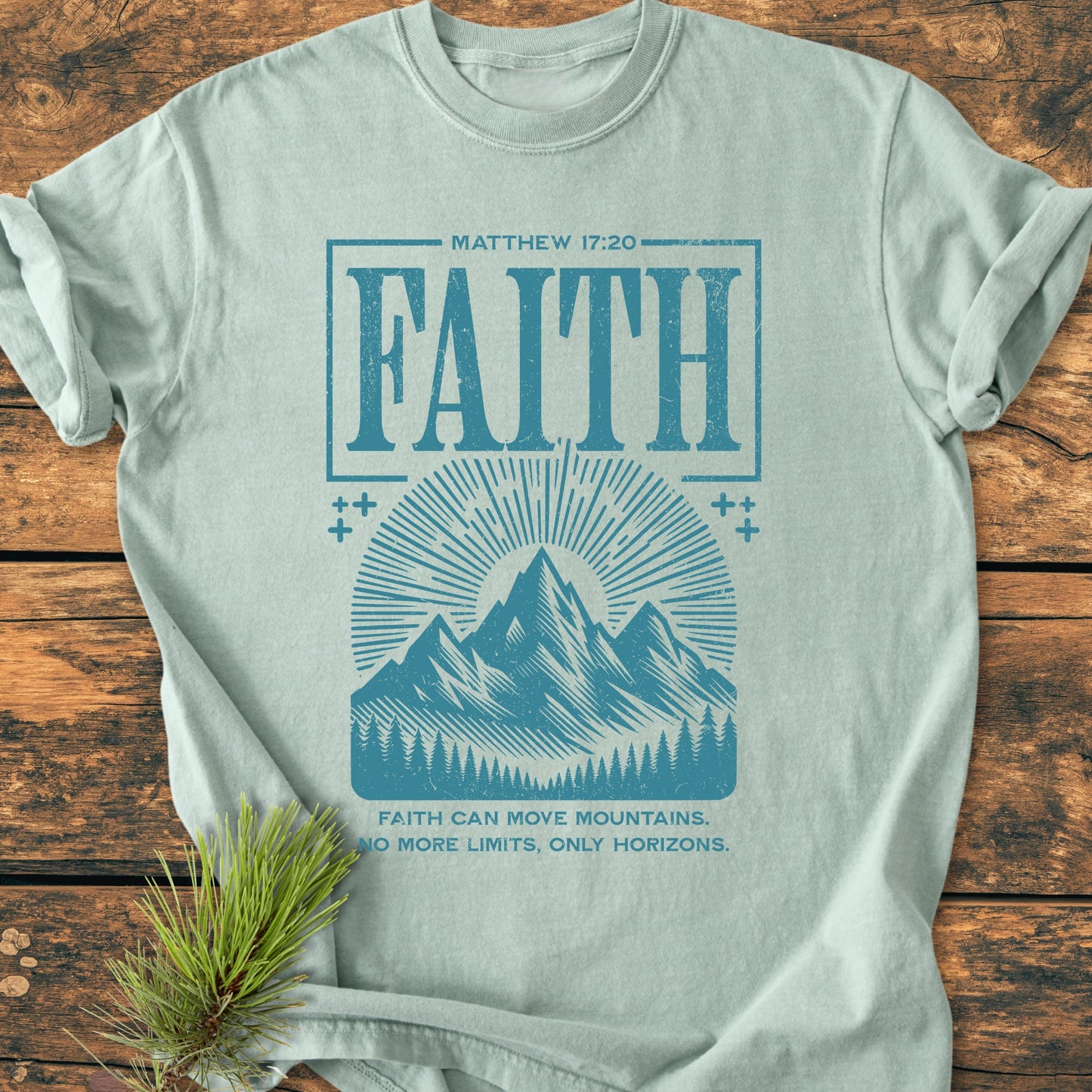 Faith Moves Mountains - Vintage Wash Tee: A seafoam T-shirt with Matthew 17:20 and FAITH in distressed letters, featuring sun rays, mountains, and pines, laid on wood with a pine sprig.