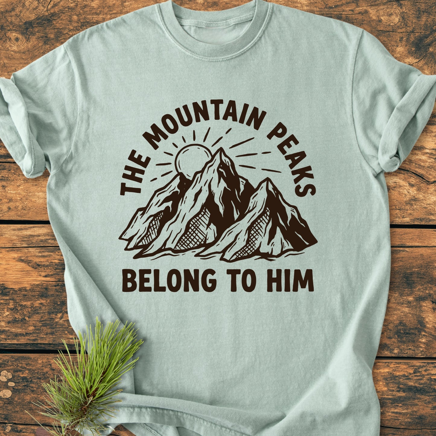 The Mountain Peaks Belong To Him - Vintage Wash Tee features a rustic mountain design with sun, in dark brown ink on a pale-green shirt, laid out with a decorative pine sprig.