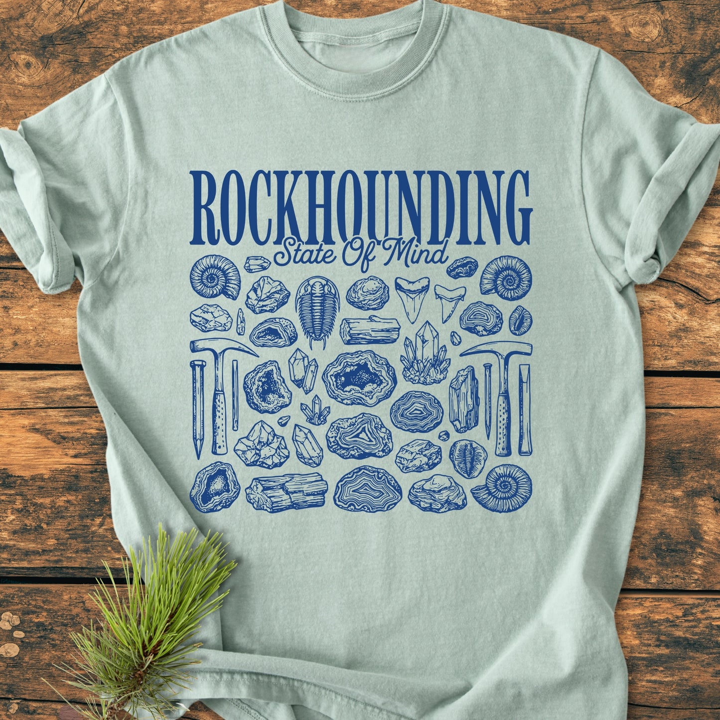 Rockhounding State Of Mind - Vintage Wash Tee featuring a bold navy graphic of rocks, fossils, and tools on light-teal fabric, laid on rustic wood with a pine sprig accent.