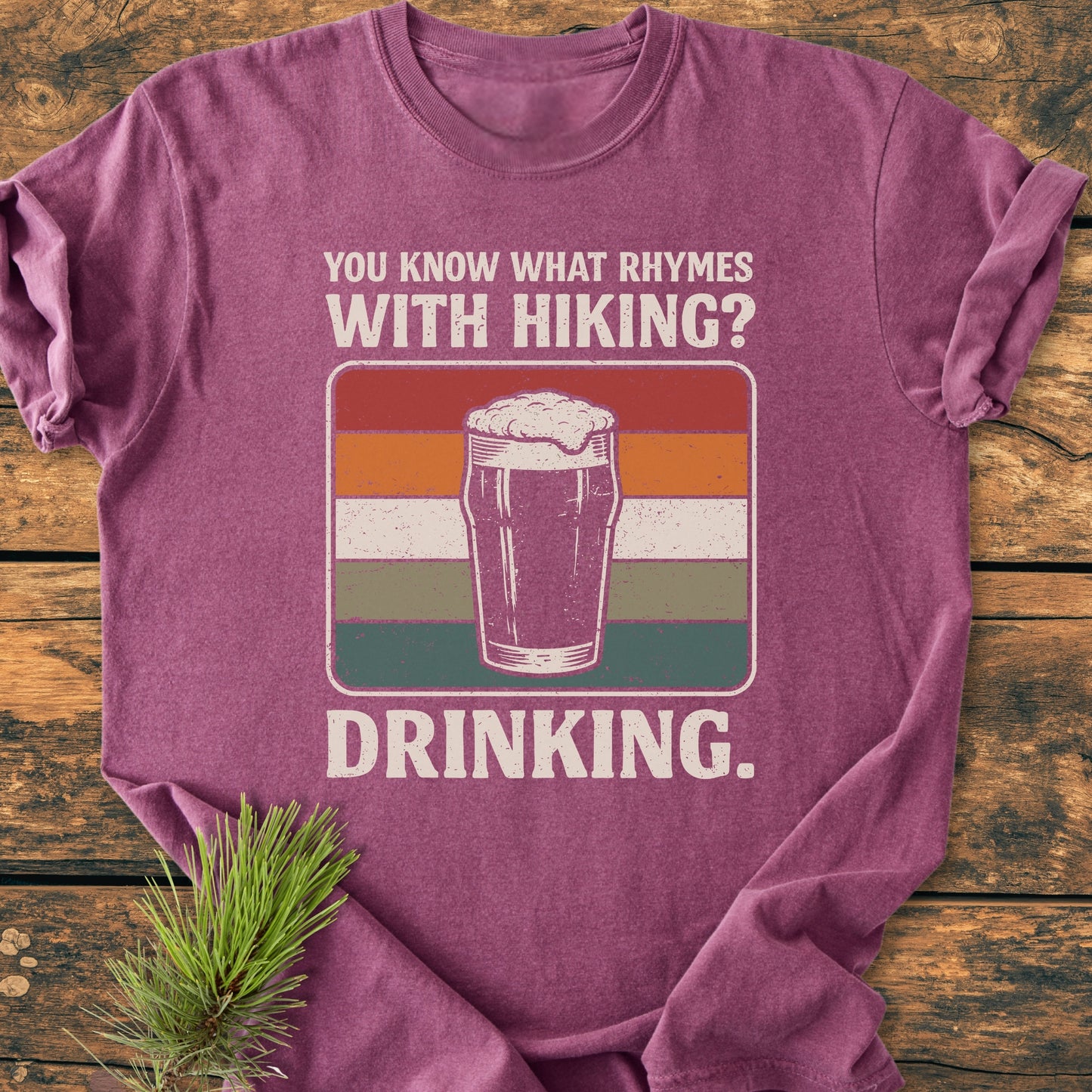 What Rhymes With Hiking? - Vintage Wash Tee featuring a humorous beer graphic and slogan, laid on wood with a pine branch, ideal for outdoor and beer enthusiasts.