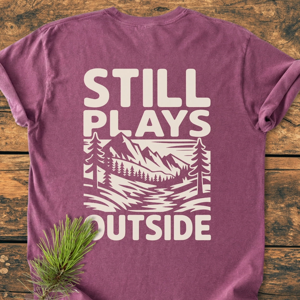 Still Plays Outside BACK PRINT - Vintage Wash Tee, featuring a stylized outdoor scene on a mauve-purple shirt, laid on wood. Pine needle sprig accents the folded garment.
