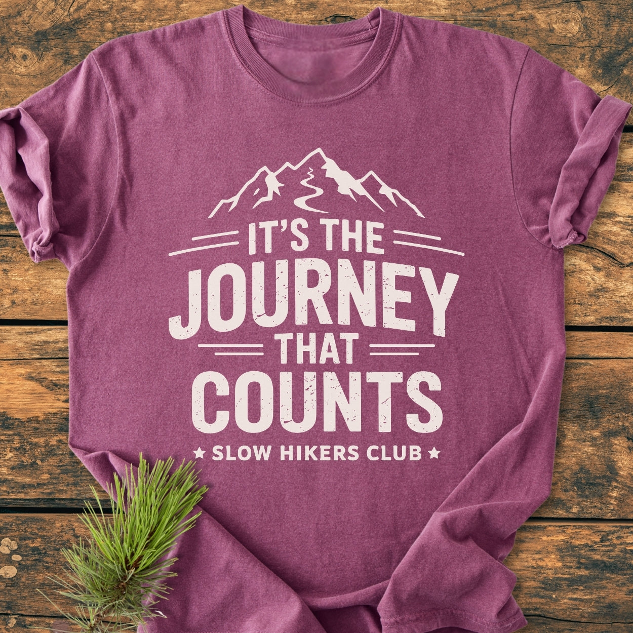 Heather-purple It's The Journey That Counts - Vintage Wash Tee features a distressed mountain graphic and Slow Hikers Club text, laid on wood with a pine sprig beside.