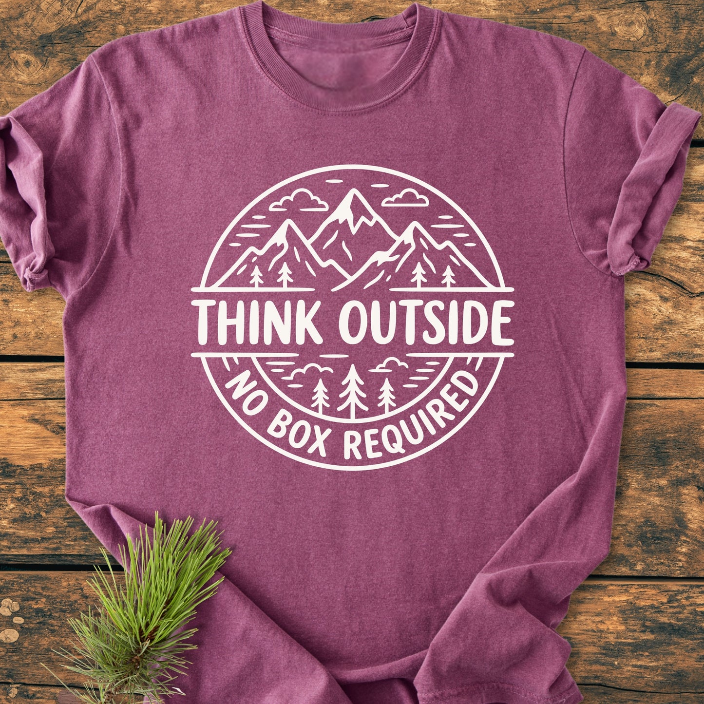Think Outside - Vintage Wash Tee features a circular graphic of mountains and trees with THINK OUTSIDE text. Laid on wood, it includes a pine sprig beside for a natural touch.