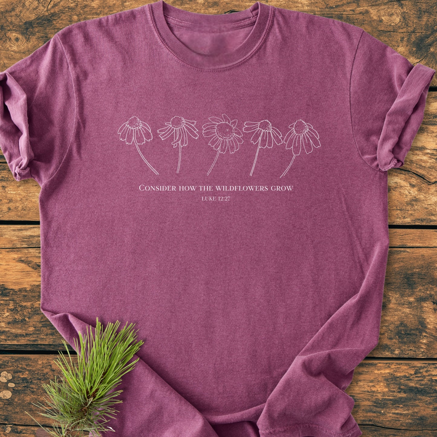Consider How The Wildflowers Grow - Vintage Wash Tee shown flat-lay, features a wildflower line drawing with Luke 12:27 inscription on a soft, heathered plum-purple fabric, styled with a pine sprig.